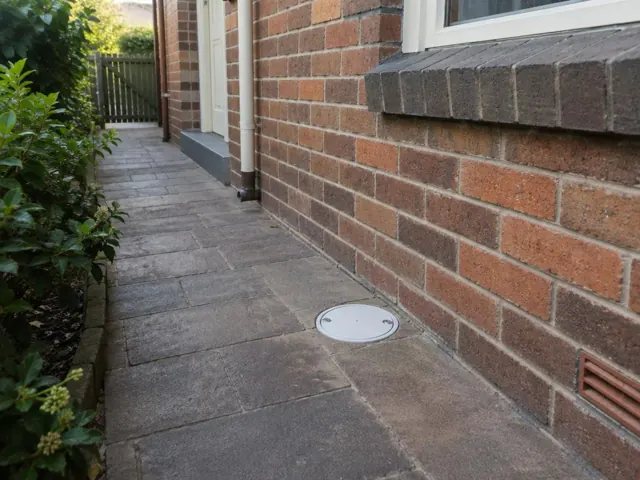 Brick side path with a round drain cover beside a house wall in Gordon